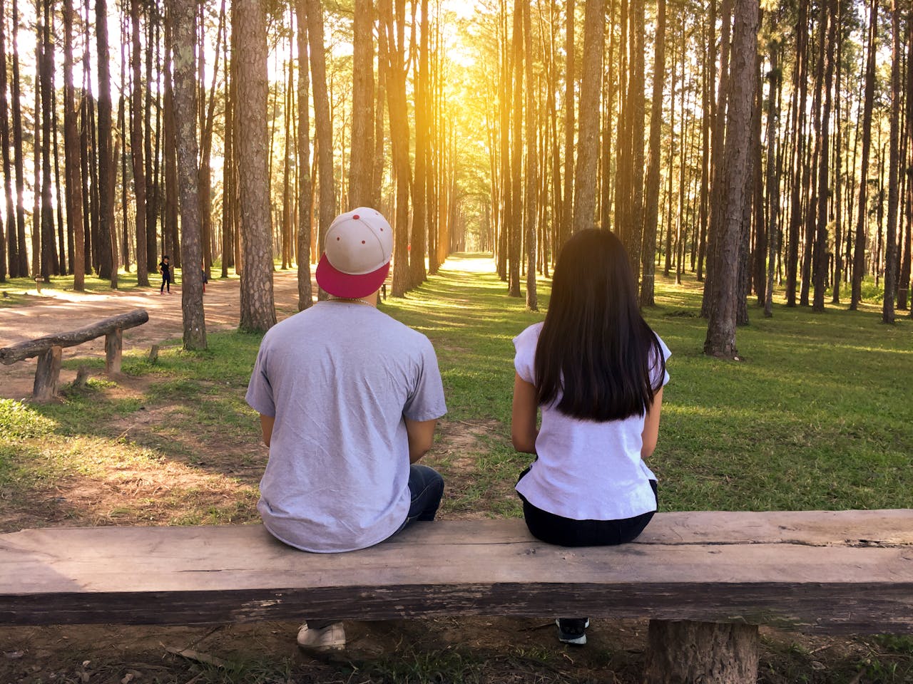 services-01 A young couple sits on a bench surrounded by tall trees in a forest at sunset.